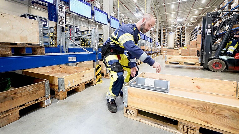 Ein Beschäftigter trägt ein Exoskelett und hebt eine Last in einem Lager an