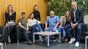 Gruppenbild: In einer Sitzecke haben sich sieben Personen versammelt. Alles lachen freundlich in die Kamera.