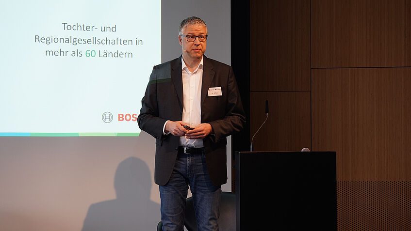 Dr. Stefan Benz (Robert Bosch GmbH) hält einen Vortrag. Im Hintergrund werden Infos gezeigt.