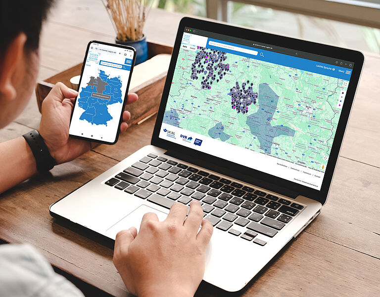 Sicher in meiner Region Junger Mann sitzt am Laptop und arbeitet mit der Anwendung "Sicher in meiner Region". Er hält ein Handy in der Hand. Die entsprechende App ist geöffnet.