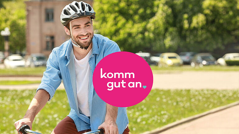 Ein lächelnder Radfahrer mit Fahrradhelm fährt durch eine Parkanlage. Im Vordergrund befindet sich ein pinker Kreis mit dem weißen Schriftzug „komm gut an“. Im Hintergrund sind Bäume, parkende Autos und ein Gebäude unscharf zu sehen.