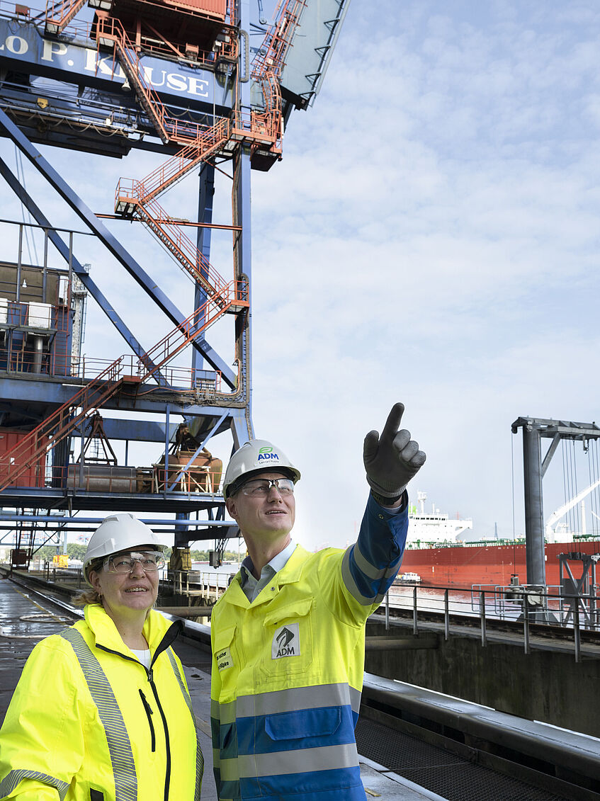 Katrin Baranowski und Lennart Rüpke, beide in gelben Warnjacken und mit weißen Schutzhelmen auf dem Kopf stehen am Terminal des Hafenbetriebs Kruse. Rüpke steht rechts von der BGHW-Mitarbeiterin und hat den linken Arm gehoben, um mit der Hand hoch auf ein Silo zu zeigen.. 