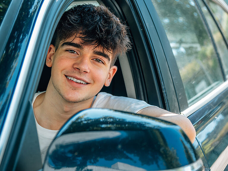 Ein junger Mann sitzt im Auto und schaut aus dem geöffneten Fenster der Fahrertür. Er lacht und sieht positiv aus.