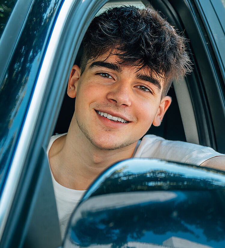 Fahranfänger 4zu3 Ein junger Mann sitzt im Auto und schaut aus dem geöffneten Fenster der Fahrertür. Er lacht und sieht positiv aus.