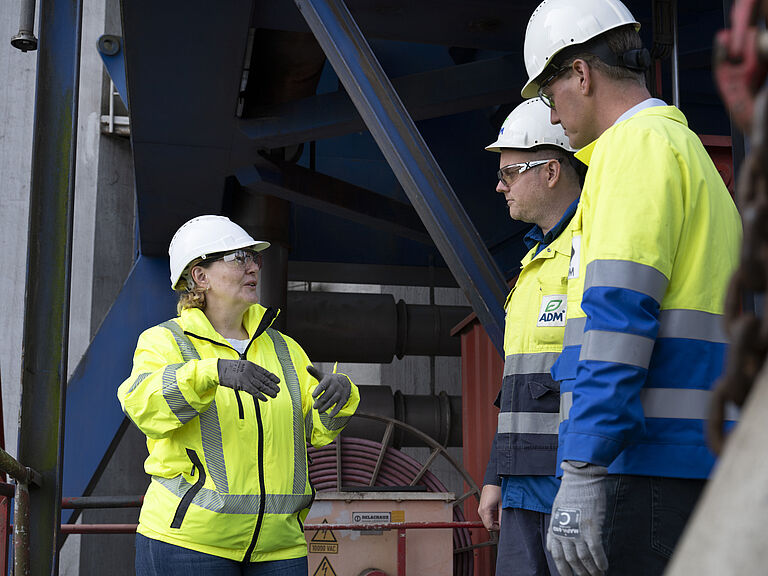 BGHW-Aufsichtsperson Katrin Baranowski spricht zu zwei Männern. Alle drei tragen gelbe Warnjacken und weiße Schutzhelme. Sie stehen auf einem Terminal bei Silo P. Kruse. 